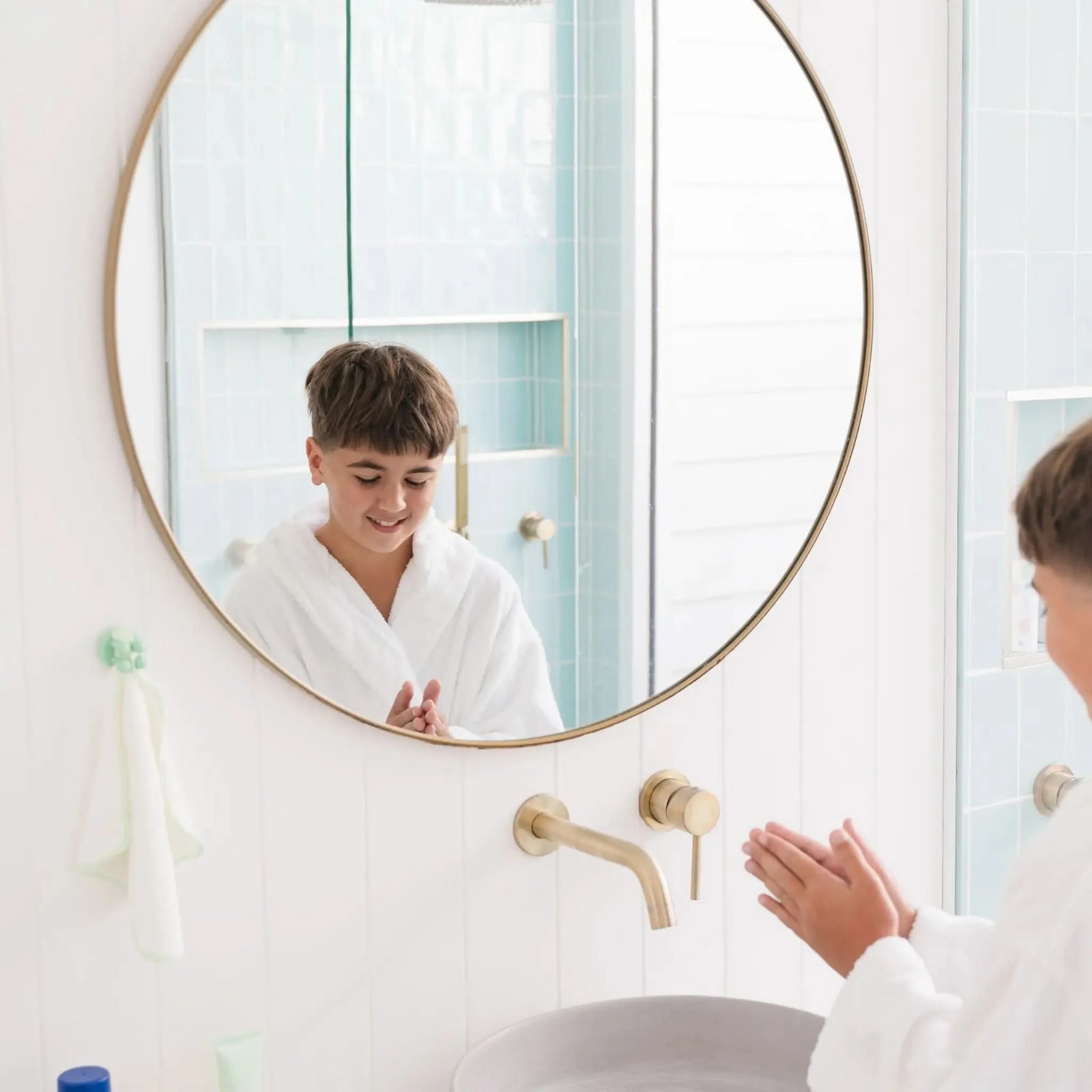 A young boy wearing a white robe is smiling and washing hands in a bright bathroom with a round mirror, where a mint colored Daily Essentials Hook is visible, Australian made and designed for ages 7 to 14. Good Skin Habits Start Early.