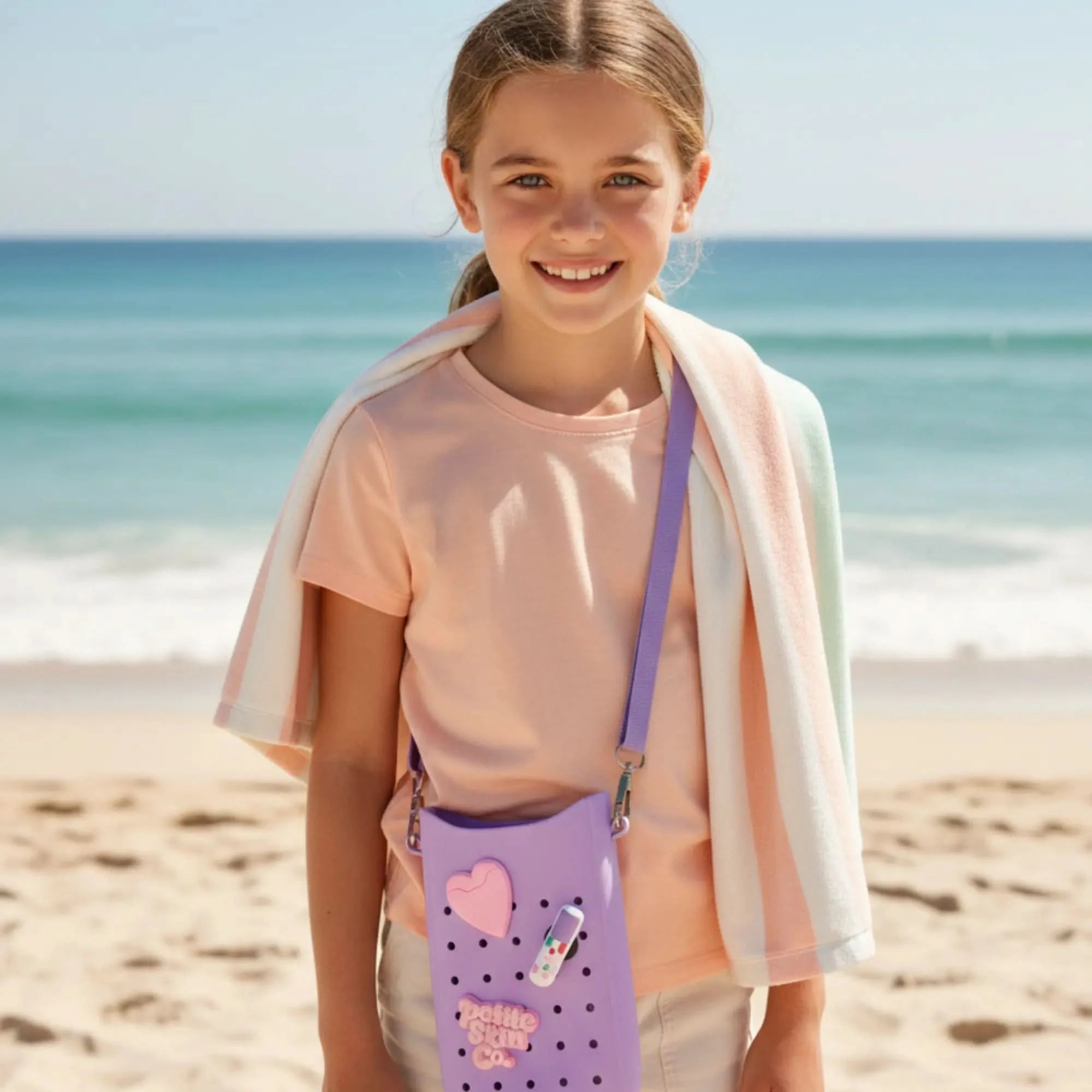 Young girl at the beach wearing a light pink shirt and a multicolor towel, carrying a Petite Crossbody Bag in lavender, Australian made, natural, designed for 7 to 14 year old skin, promoting good skin habits start early.