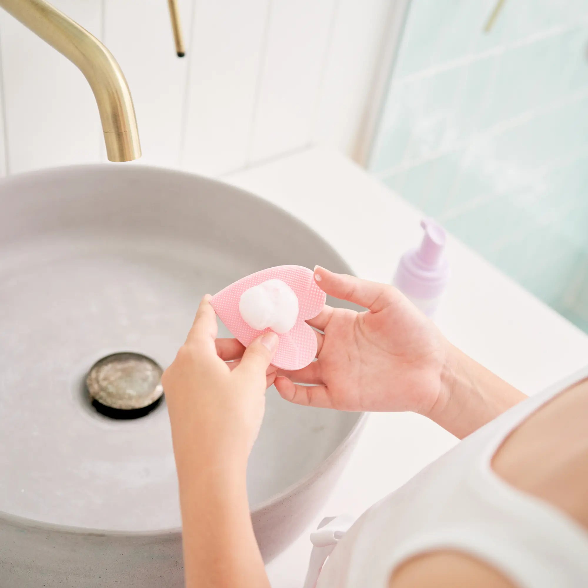 Child’s hands holding a pink silicone applicator with foam cleanser by a bathroom sink, demonstrating the Clear & Calm Skin Kit, Australian made for 7 to 14 year olds. Good Skin Habits Start Early.