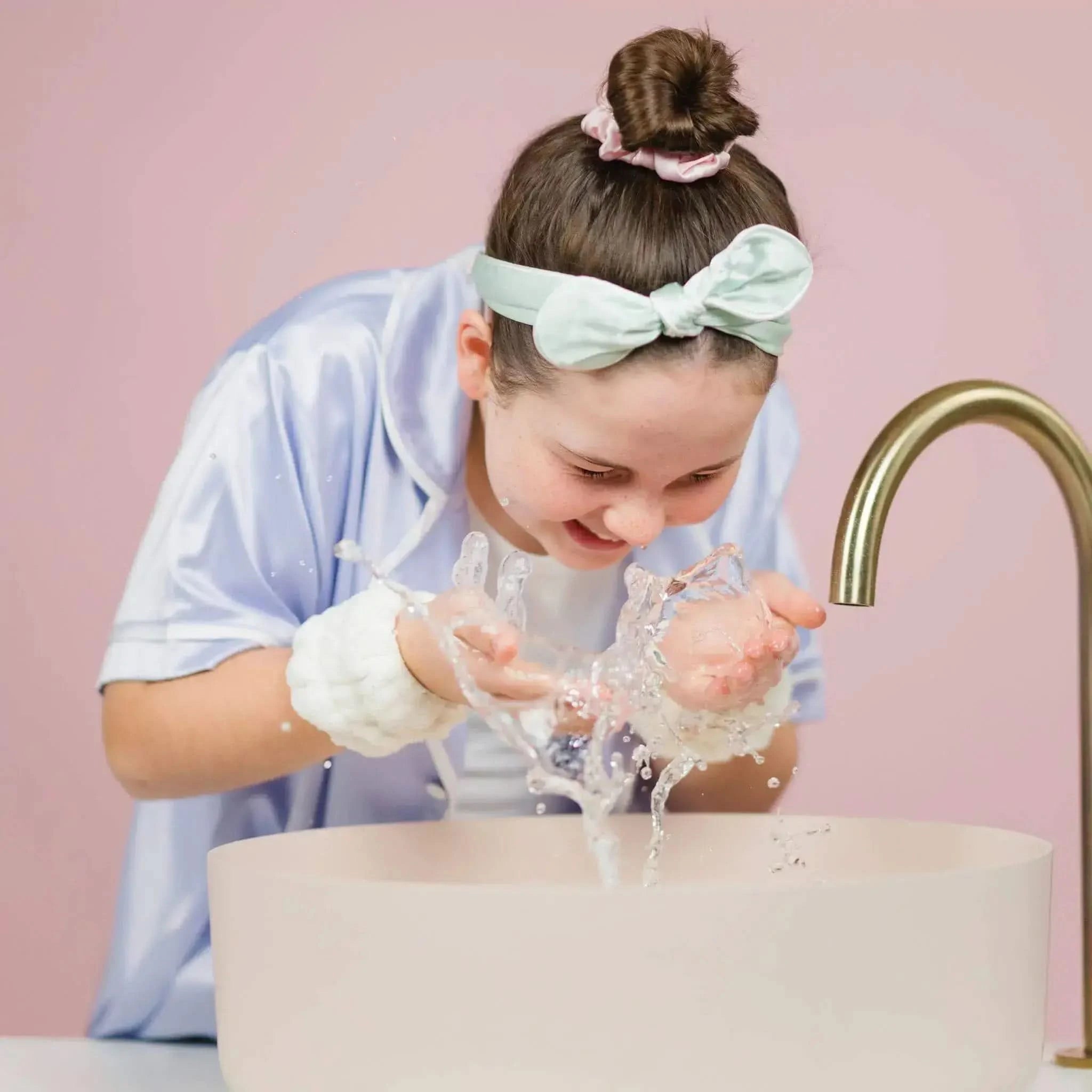A young girl wearing Dry Cuffs splashes water on her face at a sink, demonstrating gentle skincare for 7 to 14 year olds. Australian made, natural, good skin habits start early.