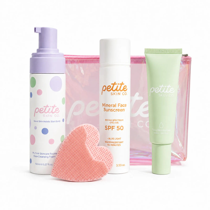 A skincare gift set from Petite Skin Co. on a white background, featuring a lavender pump bottle of facial cleansing foam, a white bottle of mineral face sunscreen with SPF 50, a light green tube of serum, a pink sponge, and a pink transparent zip pouch.