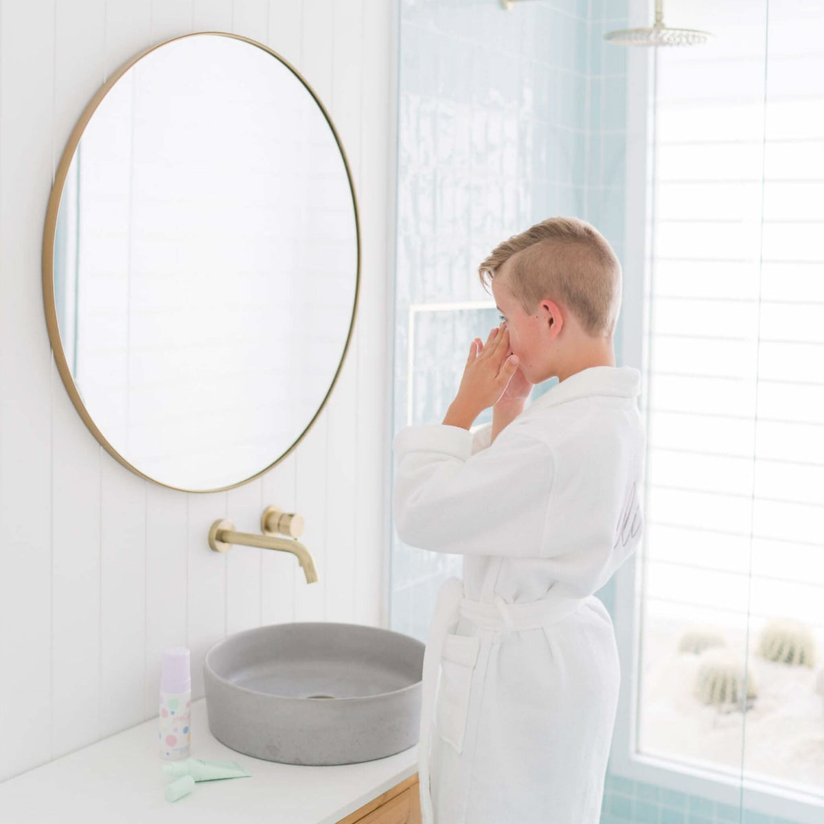 A young boy in a white bathrobe stands at a bathroom sink with a round mirror, gently touching his face in a bright, natural light setting promoting good skin habits for tweens.