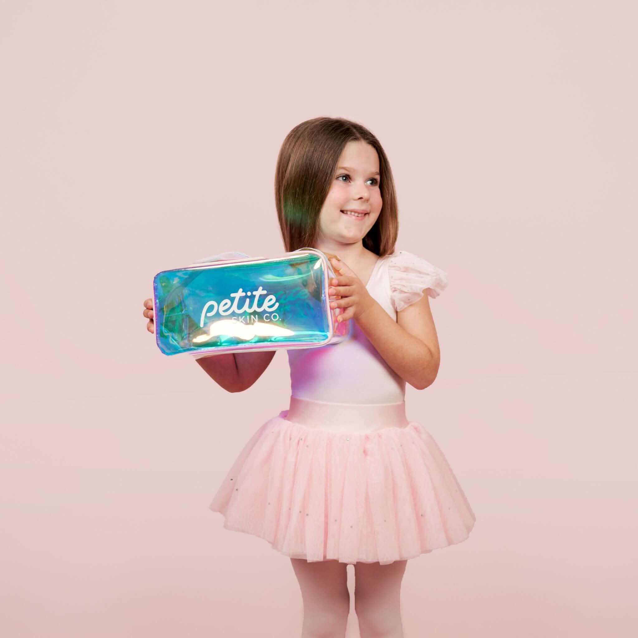 Young girl in a pink tutu dress happily holding the Holographic Skincare Bag from Petite Skin Co., featuring a roomy interior for gentle skincare products designed for 7 to 14 year olds, Australian made with natural ingredients promoting good skin habits start early.