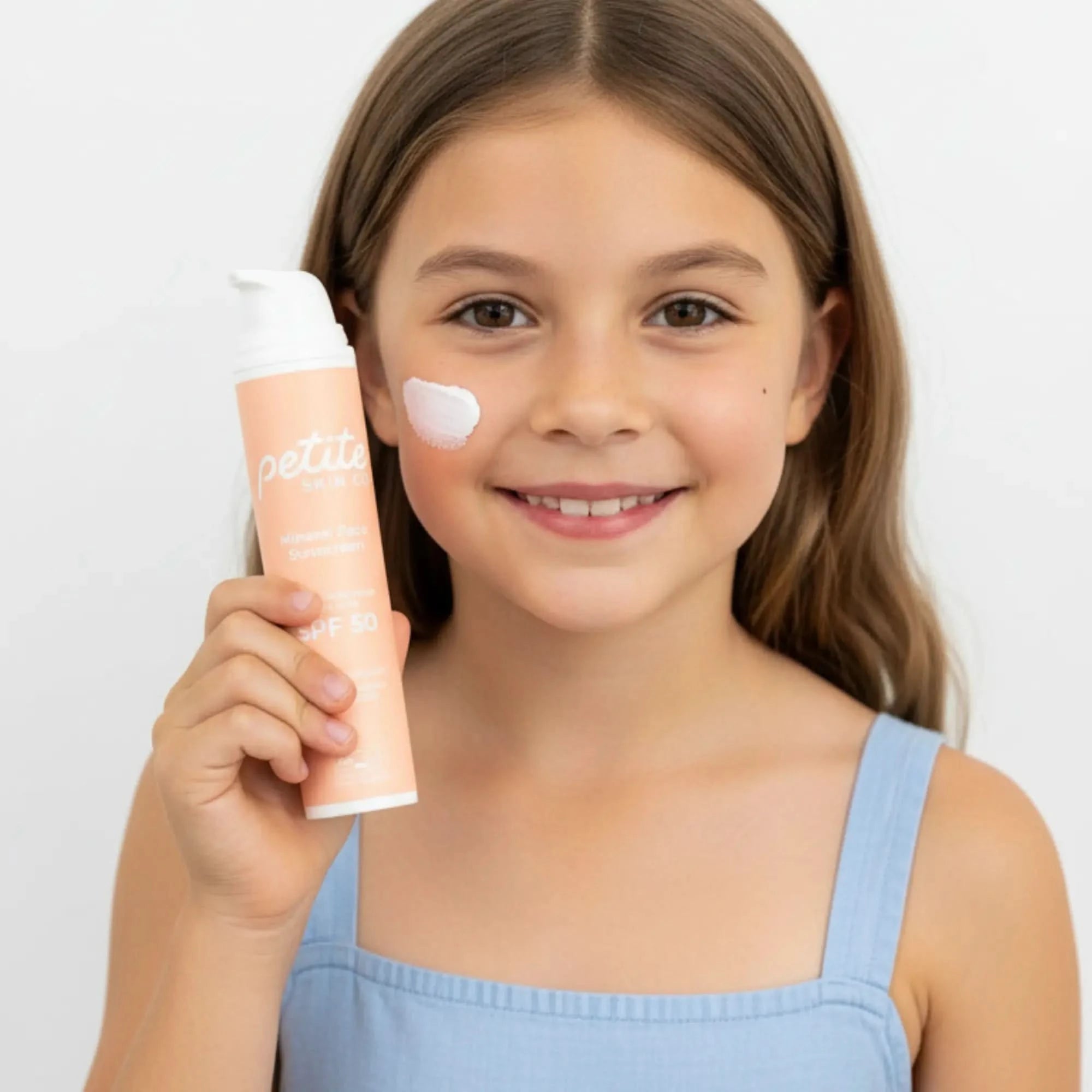 Young girl holding a petite skin co spf50 face sunscreen on her face against a plain background
