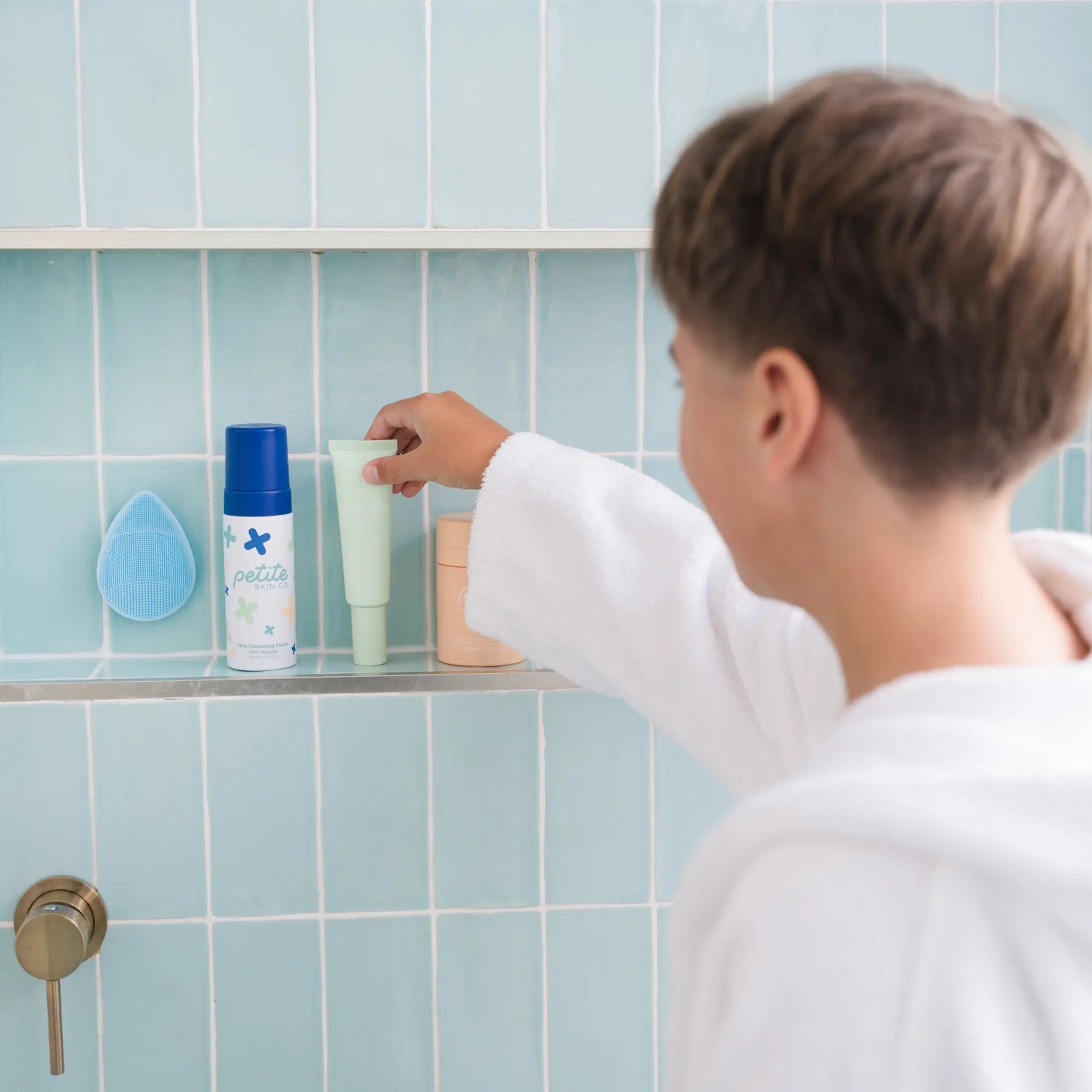 A child in a white robe reaches for a skincare tube on a bathroom shelf with blue tiles, featuring Petite Skin Co. products, highlighting the Clear Skin Essentials | Crosses Collection, Australian made and designed for 7 to 14 year olds. Good Skin Habits Start Early.