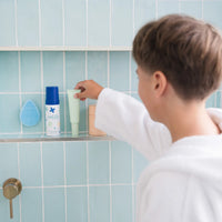 A child in a white robe reaches for a skincare tube on a bathroom shelf with blue tiles, featuring Petite Skin Co. products, highlighting the Clear Skin Essentials | Crosses Collection, Australian made and designed for 7 to 14 year olds. Good Skin Habits Start Early.