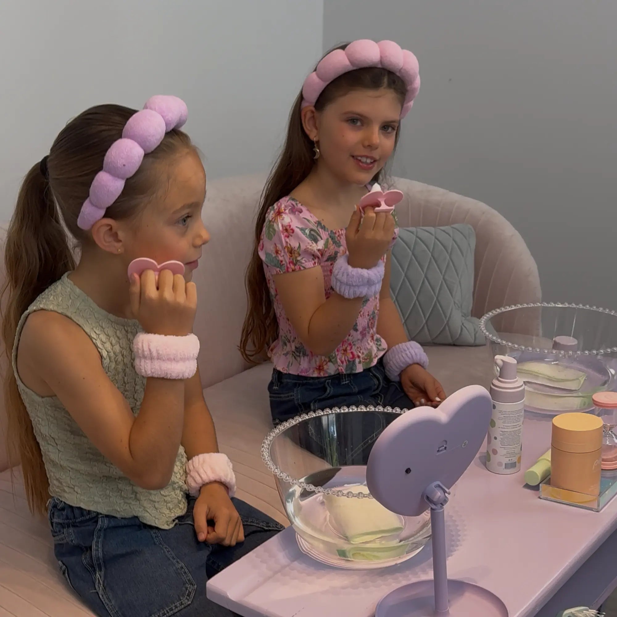 Two young girls with pink headbands and wristbands using gentle skincare products from the Skin Bliss Kit at a vanity table, demonstrating a daily skincare routine for 7 to 14 year olds.