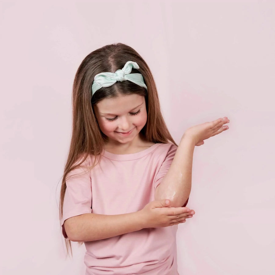 Young girl with long hair wearing a pink shirt and light green headband applying Body Cream to her elbow, showcasing Australian made, natural skincare for 7 to 14 year olds with the tagline Good Skin Habits Start Early.