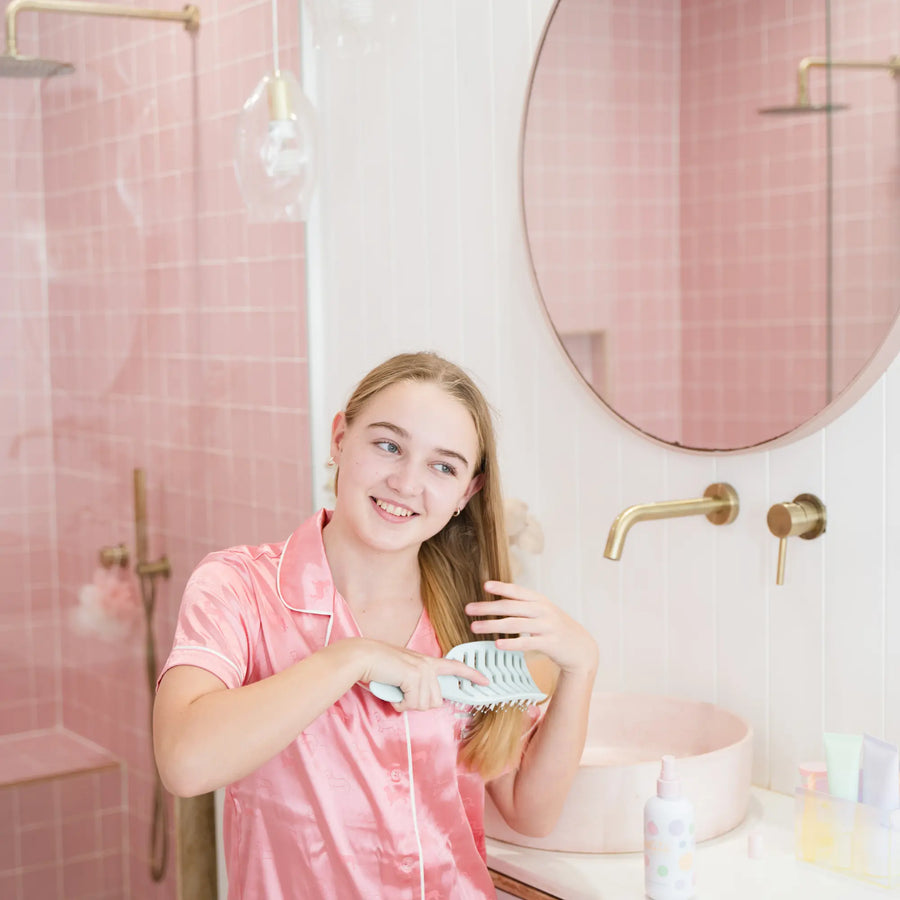 Young girl in pink satin pajamas brushing her hair with a dual-bristle brush in a pink tiled bathroom, demonstrating the Tangle-Free Duo, Australian made natural product for 7 to 14 year olds. Good Skin Habits Start Early.
