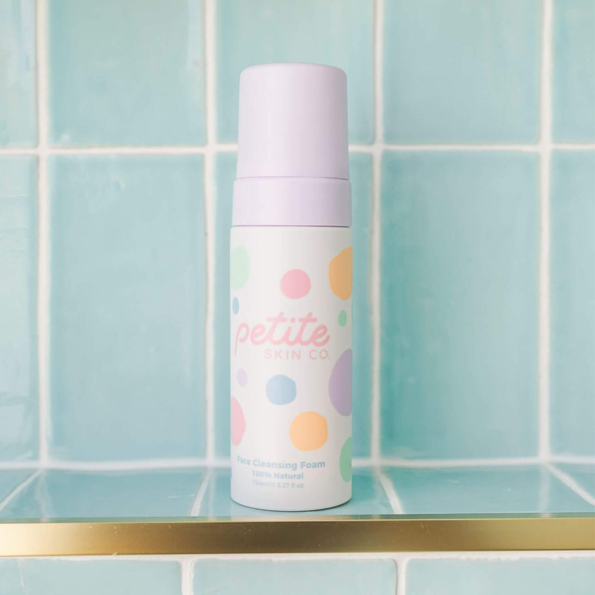 A bottle of Petite Skin Co. Face Cleansing Foam with multicolored dots design, placed against a light blue tiled background.