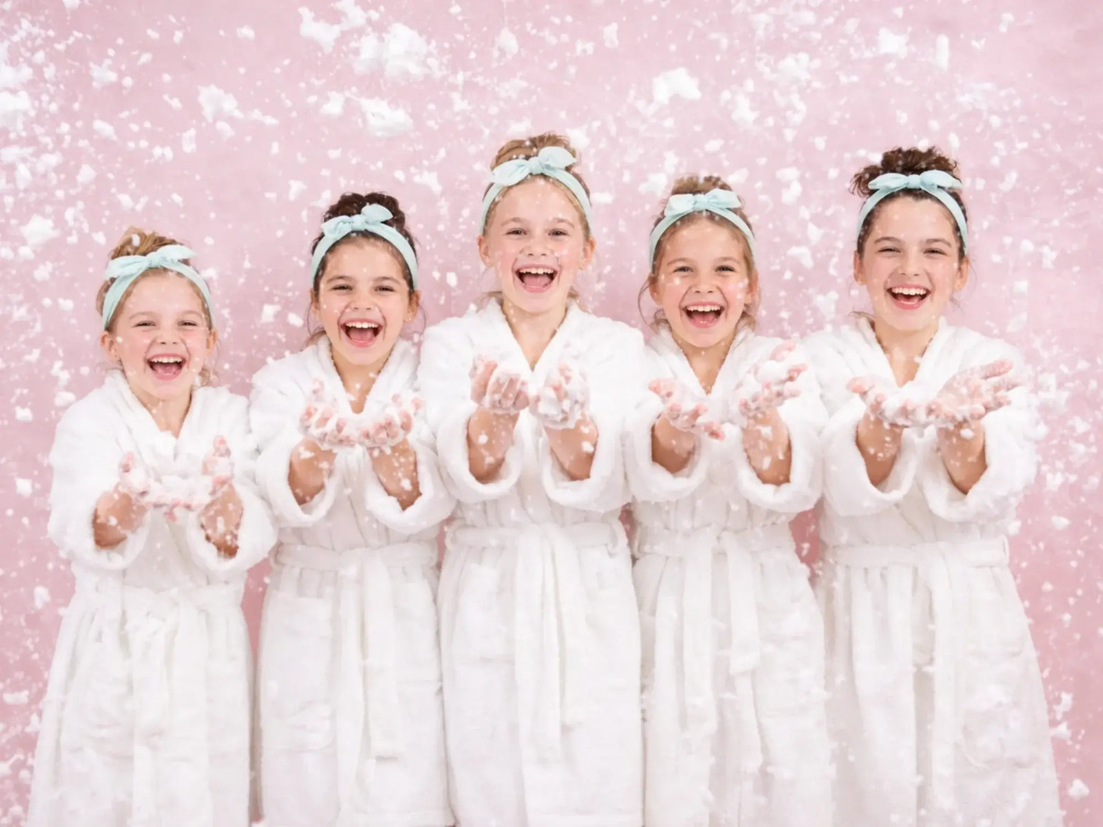 Five young girls wearing white robes and light blue headbands smiling and playing with white foam against a soft pink background, conveying joy and gentle skincare fun.