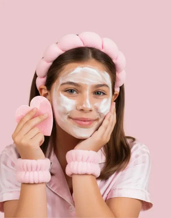 A young girl with a white face mask wears a pink flower-shaped headband, matching wrist cuffs, and a pink pajama shirt, holding a pink heart-shaped sponge against a solid pink background.