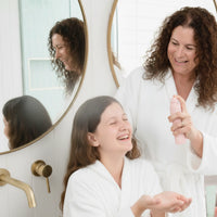 Mother and daughter in white robes smiling in a bathroom as the mother dispenses product from a pink bottle, promoting the Skin Bliss Kit with Australian made, natural skincare designed for 7 to 14 year olds – Good Skin Habits Start Early.