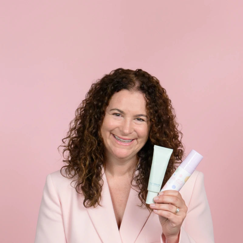 Woman with curly hair wearing a light pink blazer smiling against a pink background, holding two skincare tubes while promoting gentle skincare for young skin.
