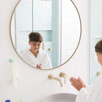 A young boy wearing a white robe is smiling and washing hands in a bright bathroom with a round mirror, where a mint colored Daily Essentials Hook is visible, Australian made and designed for ages 7 to 14. Good Skin Habits Start Early.