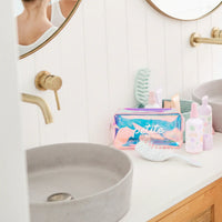 Bathroom countertop with a pink and blue iridescent toiletry bag labeled Petite alongside a white hairbrush and pastel-colored kids skincare bottles, promoting Good Hair Habits Routine. Australian made, natural, designed for the biology of 7 to 14 year old skin. Good Skin Habits Start Early.