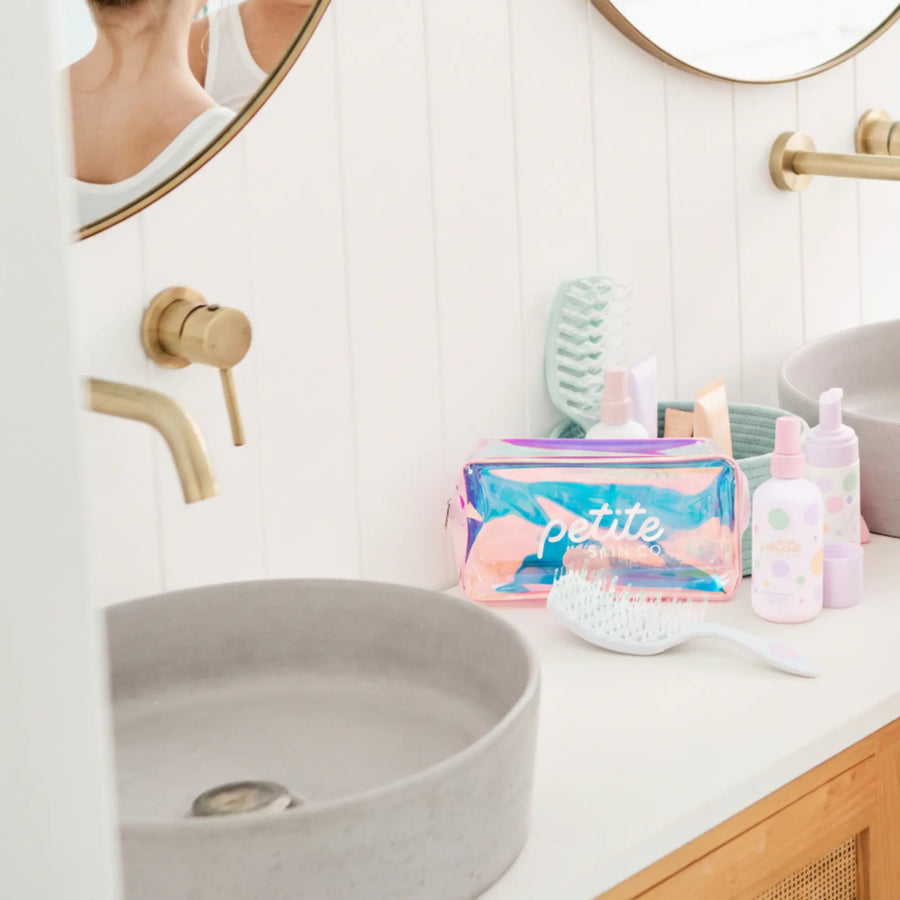 Bathroom countertop with a pink and blue iridescent toiletry bag labeled Petite alongside a white hairbrush and pastel-colored kids skincare bottles, promoting Good Hair Habits Routine. Australian made, natural, designed for the biology of 7 to 14 year old skin. Good Skin Habits Start Early.