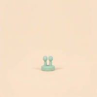 A small mint green silicone applicator brush with two rounded tips, designed to gently apply 