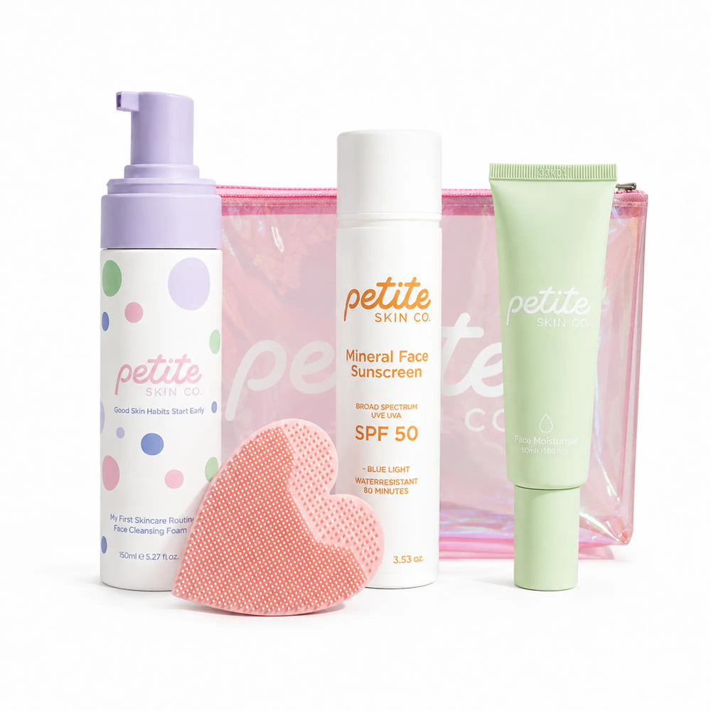 A skincare gift set from Petite Skin Co. on a white background, featuring a lavender pump bottle of facial cleansing foam, a white bottle of mineral face sunscreen with SPF 50, a light green tube of serum, a pink  sponge, and a pink transparent zip pouch.