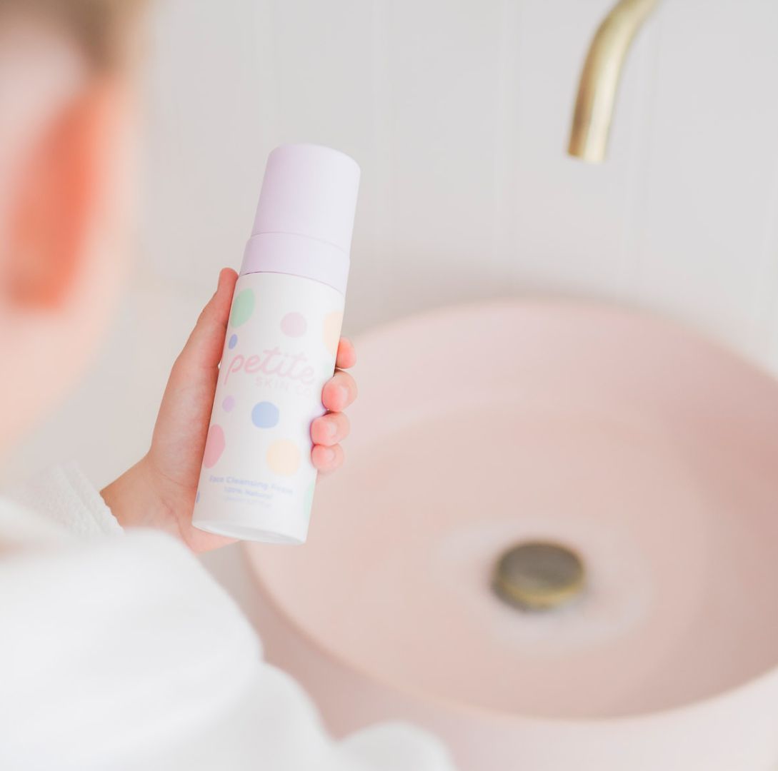 Tween girl holding petite skin co face cleansing foam and reading label over a pink bathroom sink