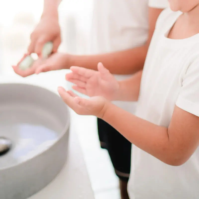 Child and adult hands near a sink with water, demonstrating a gentle face cleansing routine designed for the changing skin of 7 to 14 year olds, promoting good skin habits early.