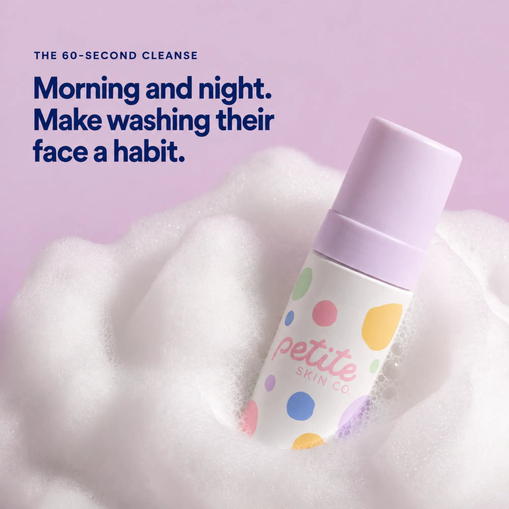 A bottle of Petite Skin Co. foaming cleanser with a lavender cap and a white body decorated with colorful polka dots, emerging from white foam against a soft lavender background with promotional text.