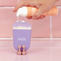 Hand holding sunscreen tube dispensing white cream onto a purple Sunscreen Applicator Brush with soft synthetic bristles, designed for even SPF application on young skin, Australian made.