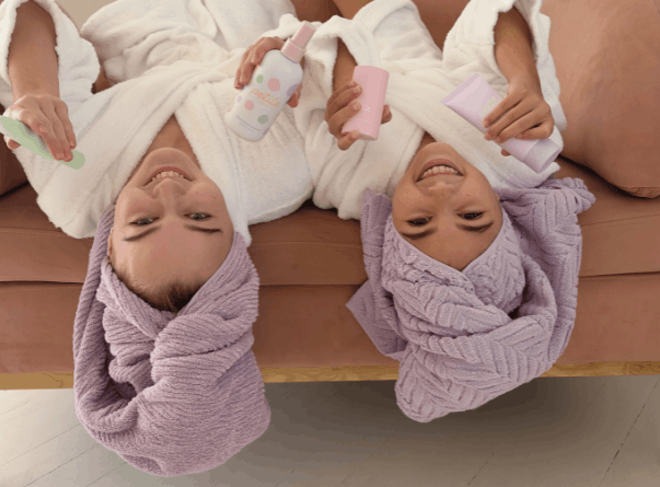 Two smiling girls with lavender towel turbans lie side by side on a couch, each holding gentle skincare bottles and tubes, showcasing a calm and natural skincare routine for young skin.