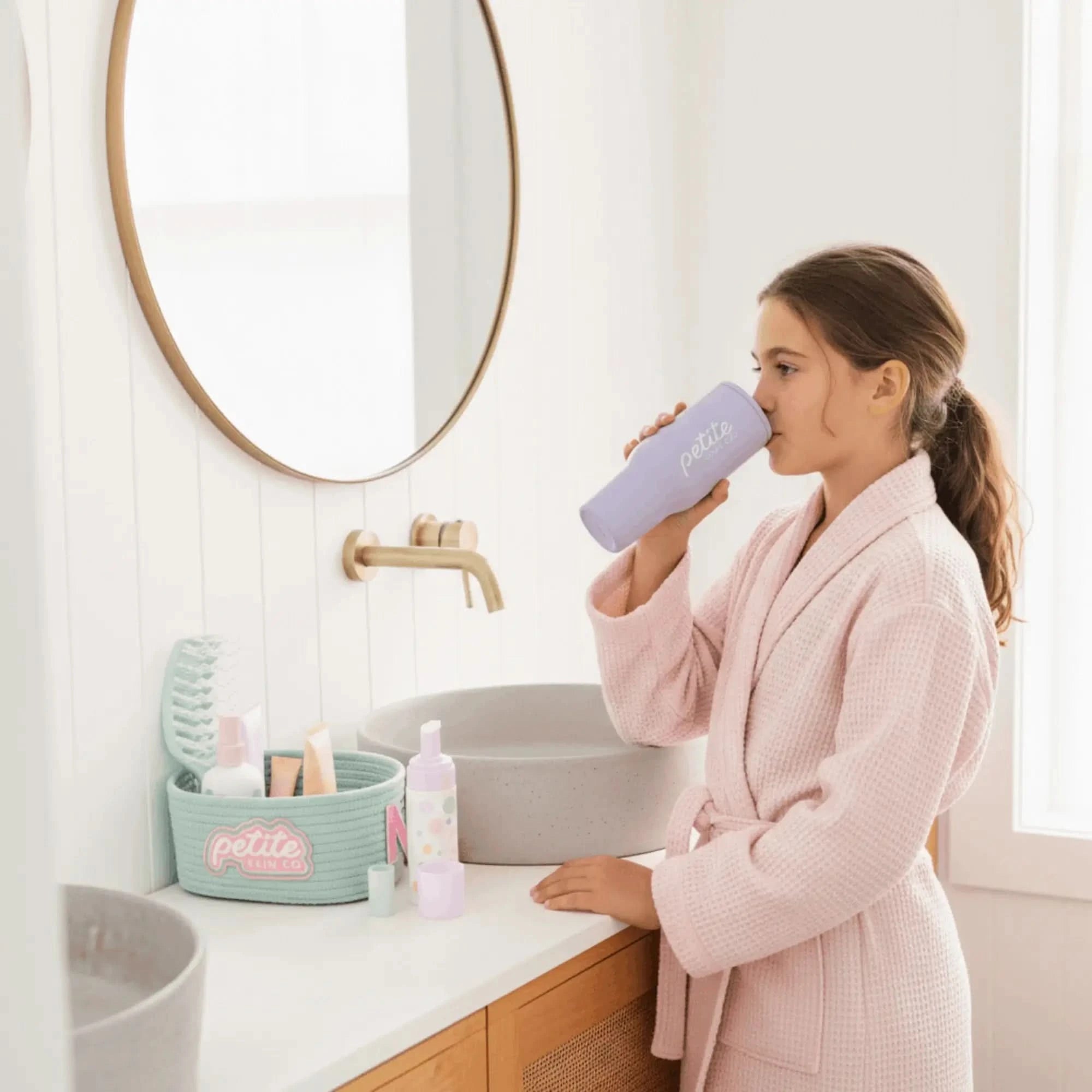 Young girl in a pink robe drinking from The Petite Hydration Cup in a bright bathroom, promoting Australian made natural skincare and good skin habits start early.